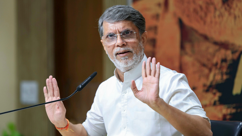 Indian man speaking at a press conference with hand raised in a stop gesture.