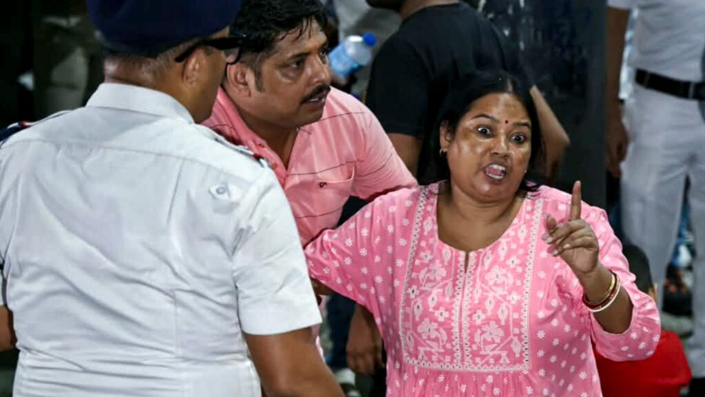 Woman in pink saree gesturing during a discussion at a crowded event.