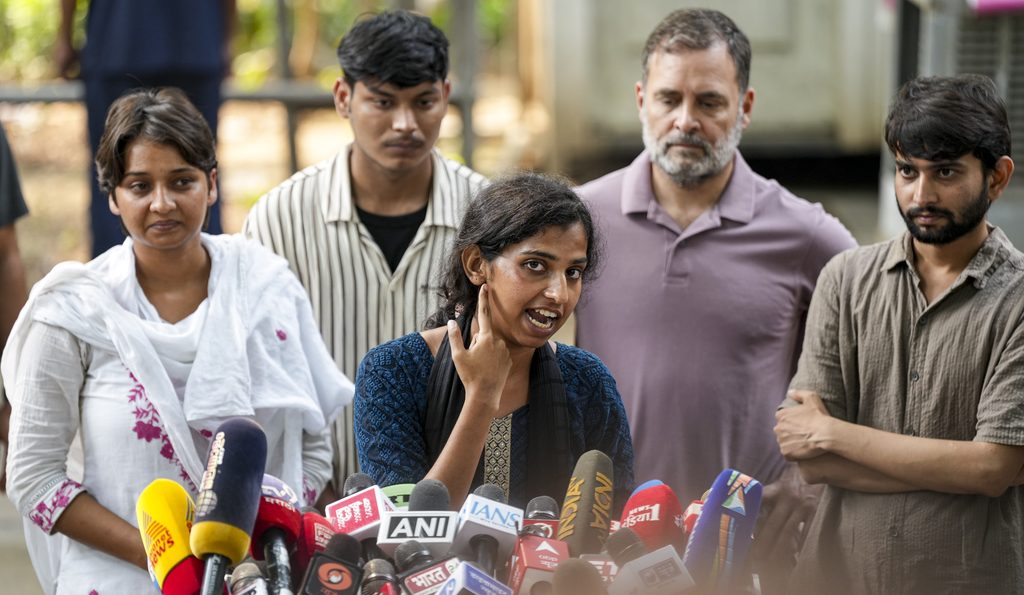 Woman speaking to media with microphones, surrounded by supporters and journalists.