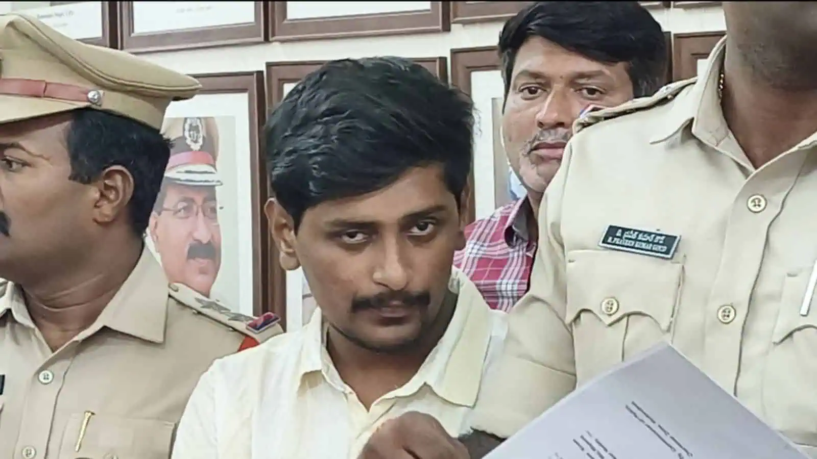 Young man with dark hair and mustache in police uniform, surrounded by officers, reading a document.