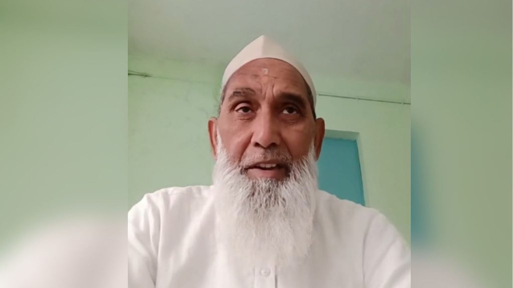 Elderly Muslim man with white beard and traditional cap indoors.